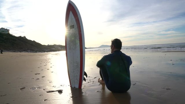 Surfer sitting on sandy beach, next to surfboard