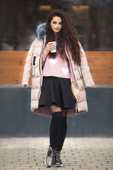 Curly hairstyle young woman alone standing near cafe and holding a cup of coffee