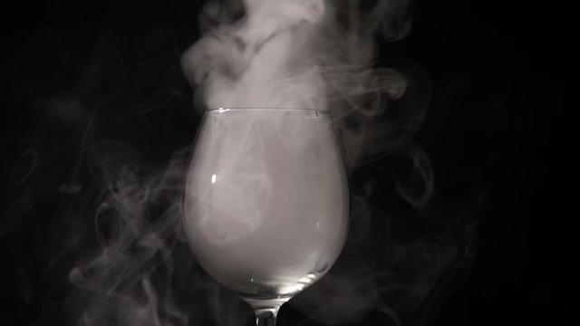 In the darkness the white smoke hovering in an empty clear glass. Cloudy is emitted from the glass vessel and dissolved slowly in the black dark. A transparent glass cup can be seen the movement of