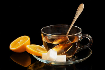 Glass cup of tea on a saucer with a lemon and sugar on the black glass background