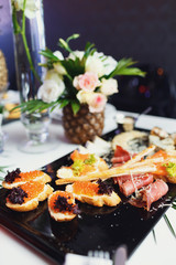 Black dish with fish snacks stands before ananas with roses