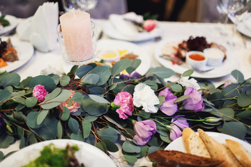 Green garland with white and violet pinks lies on dinner table