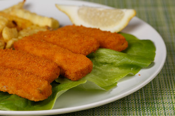 Fish fingers sticks with french fries and lemon on white plate, closeup, low focus, green canvas