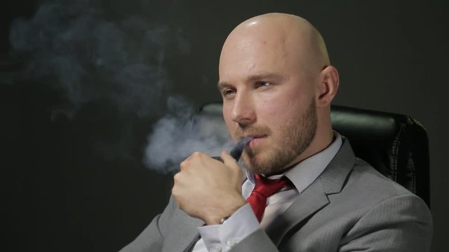 Bald man with a beard in a suit sitting on a black leather chair, looking away, thinking, lights a pipe smoking