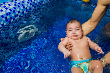 Mother teaching swimming little baby, dressed in swimmer nappies, at swimming pool, top view