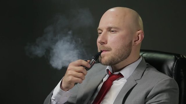 Bald man with a beard in a suit sitting on a black leather chair, looking away, thinking, lights a pipe smoking