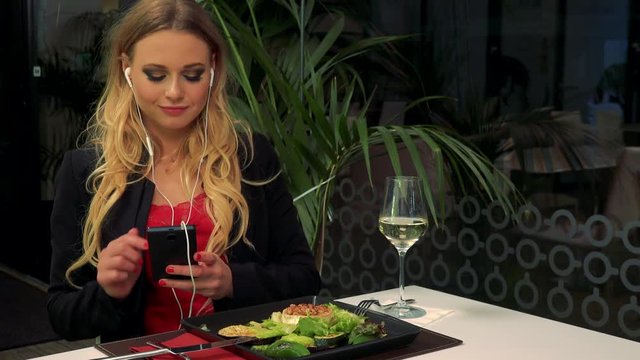 A young, beautiful woman sits at a table in a restaurant and listens to music on her smartphone, a salad and a glass of white wine in front of her