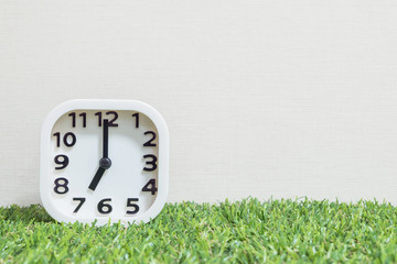Closeup white clock for decorate in 7 o'clock on green artificial grass floor and cream wallpaper textured background with copy space