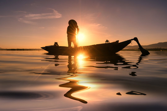 Asian Fisherman On Wooden Boat Casting A Net For Catching Freshwater Fish In Nature River In The Early Morning Before Sunrise .Silhouette Of Traditional Fishermen Throwing Net Fishing At Sunrise Time.