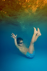 Fototapeta na wymiar A little boy is drowning, and falls into the water at the bottom of the pool on the background of bright lights and a blue background. The view from under the water. Portrait. Vertical view