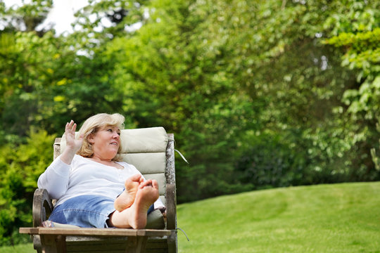 Older woman waving across the garden while sitting comfortably in a wooden chair