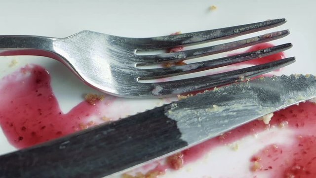 Close-up of empty white plate smeared with pink fruit syrup and crumbs, knife and fork placed on it. Remains of dessert. Detailed shot.