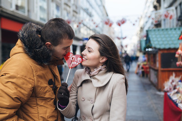 Beautiful young couple in love enjoying together at city street, kissing and holding together a lollipop in heart shape. 