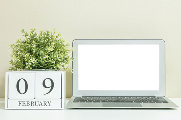 White wooden calendar with black 9 february word with white blank space at center of computer notebook on white wood desk and cream wallpaper textured background in selective focus at the calendar