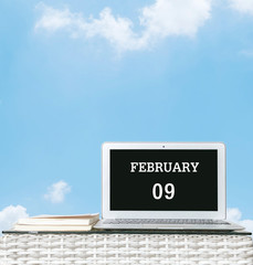 Closeup computer laptop with february 9 word on the center of screen in calendar concept on blurred wood weave table and book on blue sky with cloud textured background with copy space