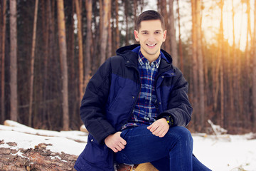 Handsome, stylish and smiling young man in jacket sitting on the woods in the forest. Early spring,...