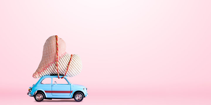 Blue retro toy car delivering craft heart for Valentine's day on pink background