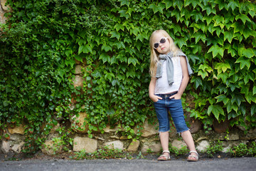 Funny little girl wearing sunglasses posing by bindweed wall on warm and sunny summer day