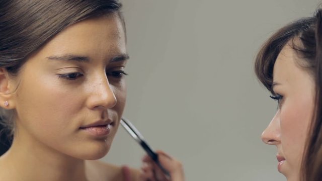 Makeup artist applying makeup with a brush on cheek of young woman. Panning
