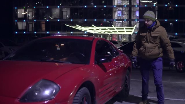 Young Man Gets Out Of A Red Car At Night At The Parking And Closes The Door. Front View.