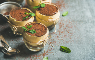 Homemade Italian dessert Tiramisu served in individual glasses with mint leaves and cocoa powder over grey concrete background, selective focus, copy space