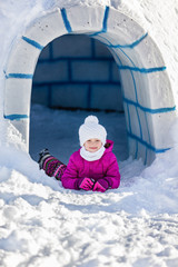 Little girl in winter clothes lying on the snow near the entrance to igloo.