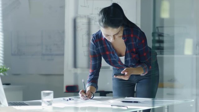 Female Design Engineer Works on Documents in a Conference Room, Last Minute Check-up, Uses Her Smartphone. In the Background Whiteboard with Schemes on it, Various Blueprints Hanging on the Walls.