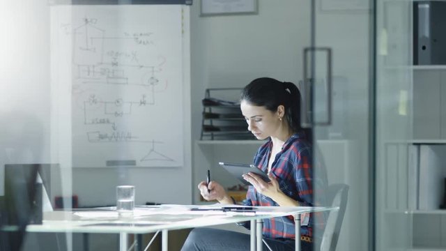 Female Design Engineer Sits at the Glass Table in Her Office, Works on a Tablet Computer, Blueprints Laying on Her Desk. Colleague Comes in. In the Background Whiteboard with Diagrams and Sketches. 