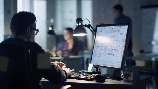 Late at Night in the Office. Design Engineer Works on His Personal Computer. On His Display We See Blueprints. Office Looks Modern. In the Background People Working.  