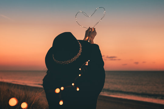 San Valentino. Cuore luminoso tenuto in mano da una ragazza vestita di nera sulla spiaggia al tramonto.