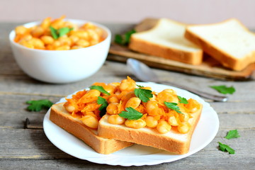 Open sandwiches with baked white beans. Beans baked with carrots, garlic and tomato sauce on a bowl, bread slices, spoon, fresh parsley on a rustic wooden table. Vegetarian and vegan protein dish