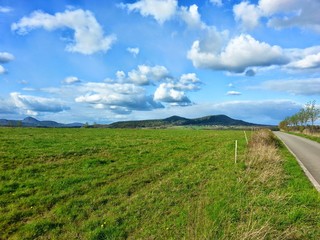 Fototapeta premium The blooming meadow with nice blue sky with dispersed clouds by sunny day. The Czech nature. Hills in the background.