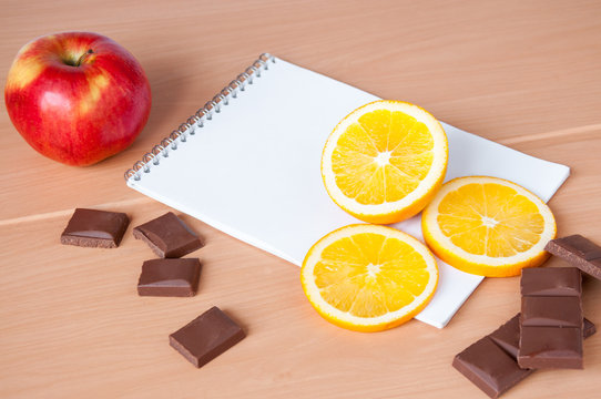 Slices Of Orange, Chocolate And Red Apple Lying On The Notepad