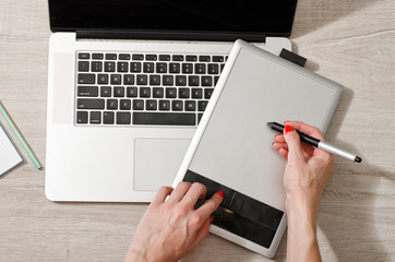 Female hand with a stylus on a graphic tablet, laptop open on a light table, top view