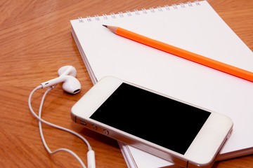 Perspective view of cellphone earphones and notebook with pencil on top of it.