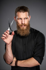 Serious hipster man with beard and mustashes holding scissors, over grey background