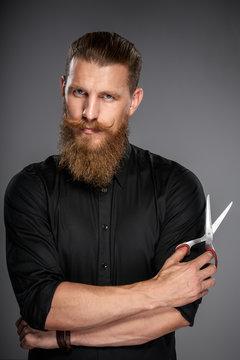 Serious hipster man with beard and mustashes holding scissors, over grey background