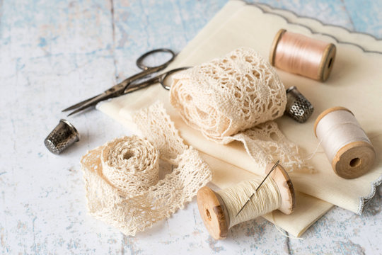 
 Sewing supplies and accessories for needlework.
 Fabric, openwork lace, spools of thread, scissors and thimbles on an old wooden table.