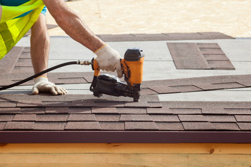 Construction worker putting the asphalt roofing (shingles) with nail gun on a new frame house - obrazy, fototapety, plakaty
