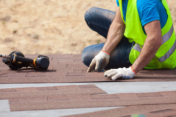 Roofer builder worker with nailgun installing Asphalt Shingles or Bitumen Tiles on a new house under construction