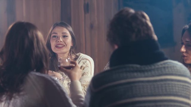 Group of happy young people laugh and chat at dinner table in a rustic cabin. Friendship, relax at holidays and week-end. Men and women drinking mulled wine. 4K UHD 60 FPS RAW edited footage