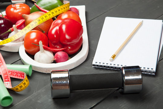 Sport and diet. Healthy lifestyle. Vegetables, dumbbells  notebook. Peppers, tomatoes, garlic, onion  radish in a heart on  black background