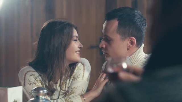 Group of happy young people laugh and chat at dinner table in a rustic cabin. Friendship, relax at holidays and week-end. Guy kisses a girl. 4K UHD 60 FPS RAW edited footage