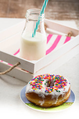 A rainbow sprinkled donut 'doughnut on a colorful plate. A white wooden crate with a milk bottle in the background. 