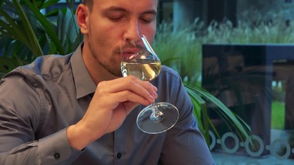 A young, handsome man sits at a table in a restaurant, takes a sip of wine and looks around