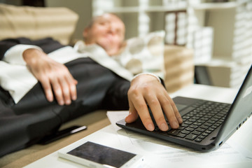 Businessman in suit lying on a couch with two cellphones and laptops, sleeping. Exhausted man early morning hand on keyboard,. Responsible executive working fell asleep. 