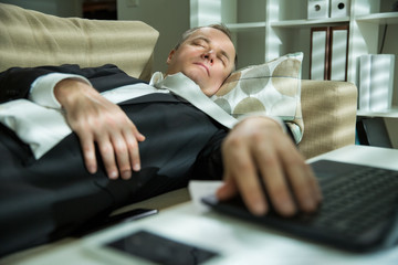 Businessman in suit lying on a couch with two cellphones and laptops, sleeping. Exhausted man early morning hand on keyboard,. Responsible executive working fell asleep. 