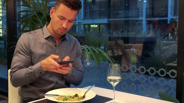 A young, handsome man sits at a table in a restaurant and takes a picture of his meal with a smartphone