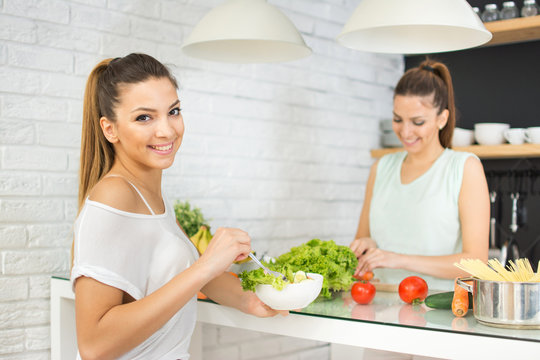 Young woman eating fresh salad. Her sister cutting vegetables in the background.