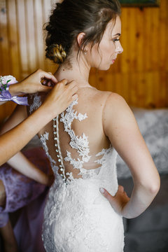 Bride holds her hands on waist while lady buttons up her lace dr